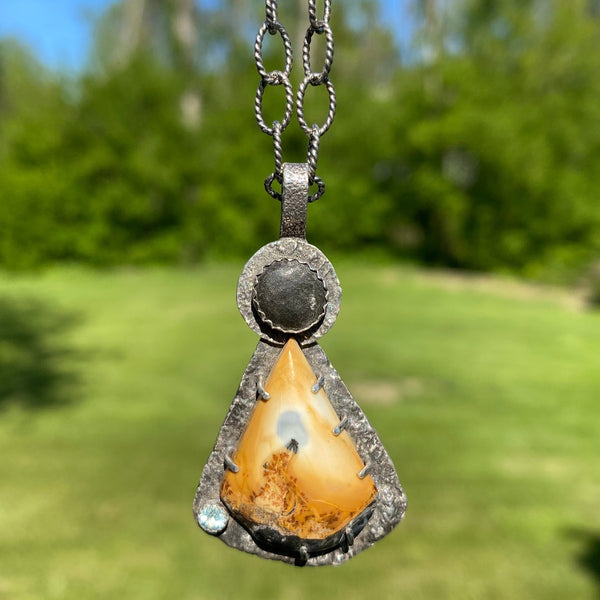 The focal point of this three stone charm is a beautifully patterned Sonoran Dendritic Jasper.  Mostly golden yellow with bits of black, deep orange and sky blue, it brings to mind a volcanic fire rising to the sky.  A round, black beach stone and faceted Sky Blue Topaz complete the trio.
Held in place with prongs, the Sonoran Dendritic is an irregularly shaped triangular stone with a curved bottom and measures 1" in length and .75" in width.  An 8mm Lake Michigan beach stone tops the piece
