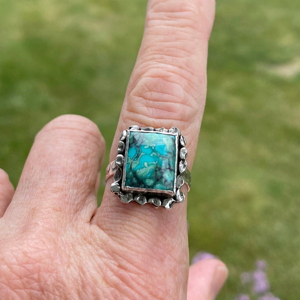 Hidden in the beauty of the blue, green and black patterned Turquoise is a tiny green heart.  Do you see it?
Created by hammering and playing with fire, a randomly textured border surrounds the rectangular stone.  Further hammer-play created the circular pattern on the bezel, backplate and ring shank. 
Made with Sterling Silver and a 12x10mm Hubei Turquoise.
Size 7.75
P.S.  It's in the upper right hand corner.  Did you know green is the color of the Heart Chakra?