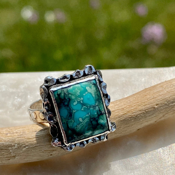 Hidden in the beauty of the blue, green and black patterned Turquoise is a tiny green heart.  Do you see it?
Created by hammering and playing with fire, a randomly textured border surrounds the rectangular stone.  Further hammer-play created the circular pattern on the bezel, backplate and ring shank. 
Made with Sterling Silver and a 12x10mm Hubei Turquoise.
Size 7.75
P.S.  It's in the upper right hand corner.  Did you know green is the color of the Heart Chakra?