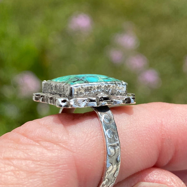 Hidden in the beauty of the blue, green and black patterned Turquoise is a tiny green heart.  Do you see it?
Created by hammering and playing with fire, a randomly textured border surrounds the rectangular stone.  Further hammer-play created the circular pattern on the bezel, backplate and ring shank. 
Made with Sterling Silver and a 12x10mm Hubei Turquoise.
Size 7.75
P.S.  It's in the upper right hand corner.  Did you know green is the color of the Heart Chakra?