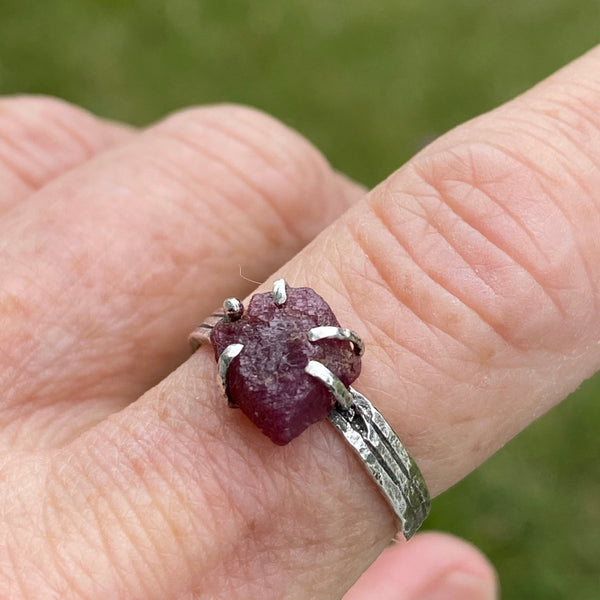 Five Sterling Silver prongs hold a raw Ruby on a textured Sterling Silver band.    Worn by itself or as part of a stack, this July birthstone stands out with its unique shape.  The flat topped Ruby with irregular edges measures 8.80mm x 7.70mm across at it widest points.  It is 3.50mm deep.  Ruby is said to stimulate the Heart Chakra offering nurturing and spiritual wisdom.  Size 7.75